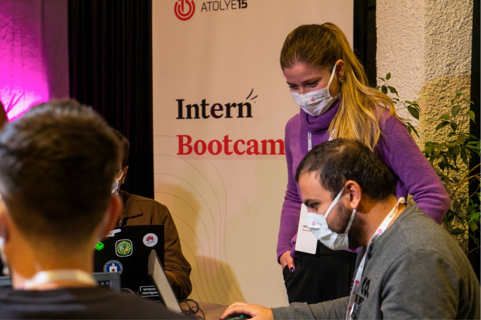 A man and a blond girl in a purple sweater, wearing face masks in front of a banner that says “Atolye15 Intern Bootcamp."