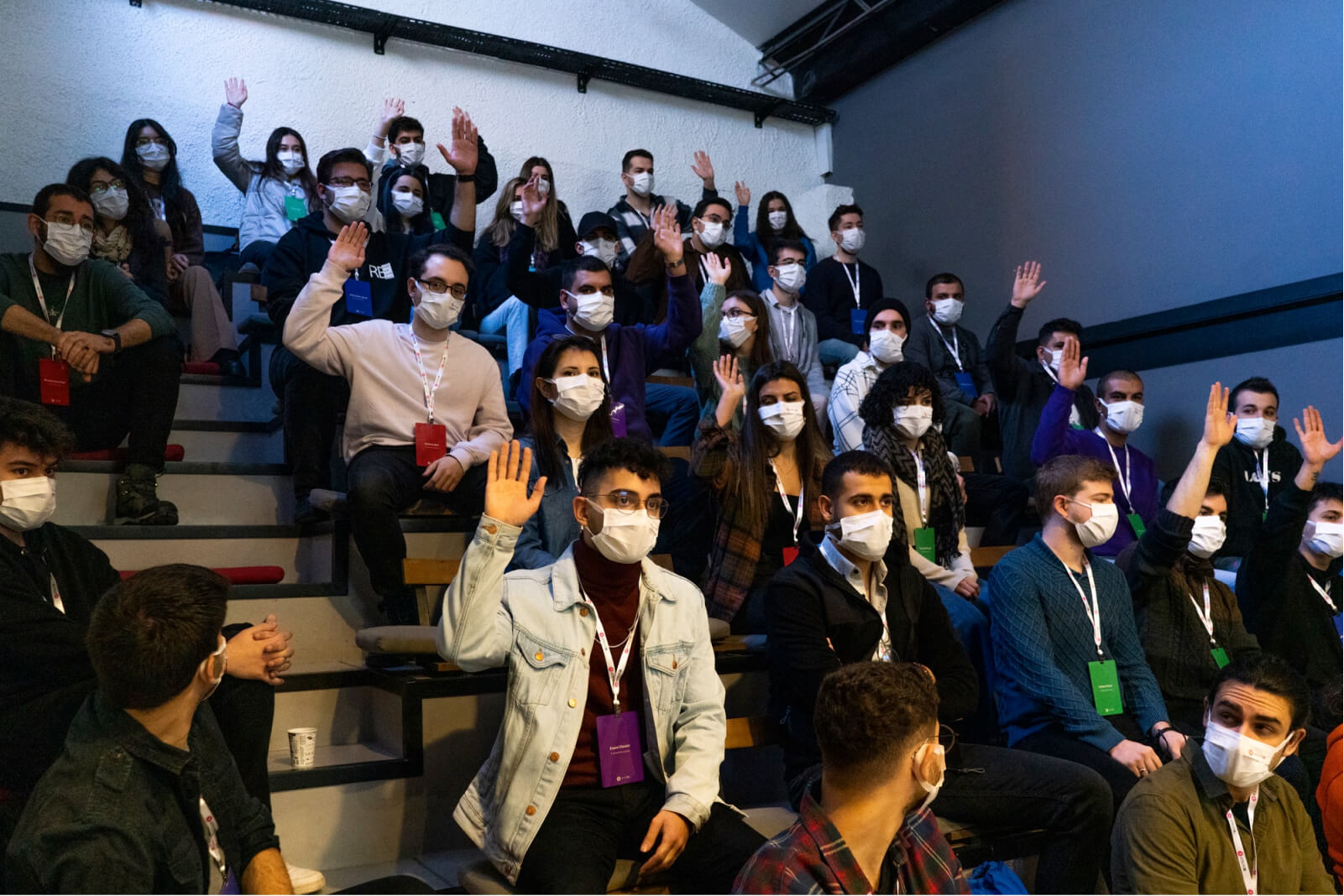 A group of young people in a lecture hall wearing face masks and raising their hands.