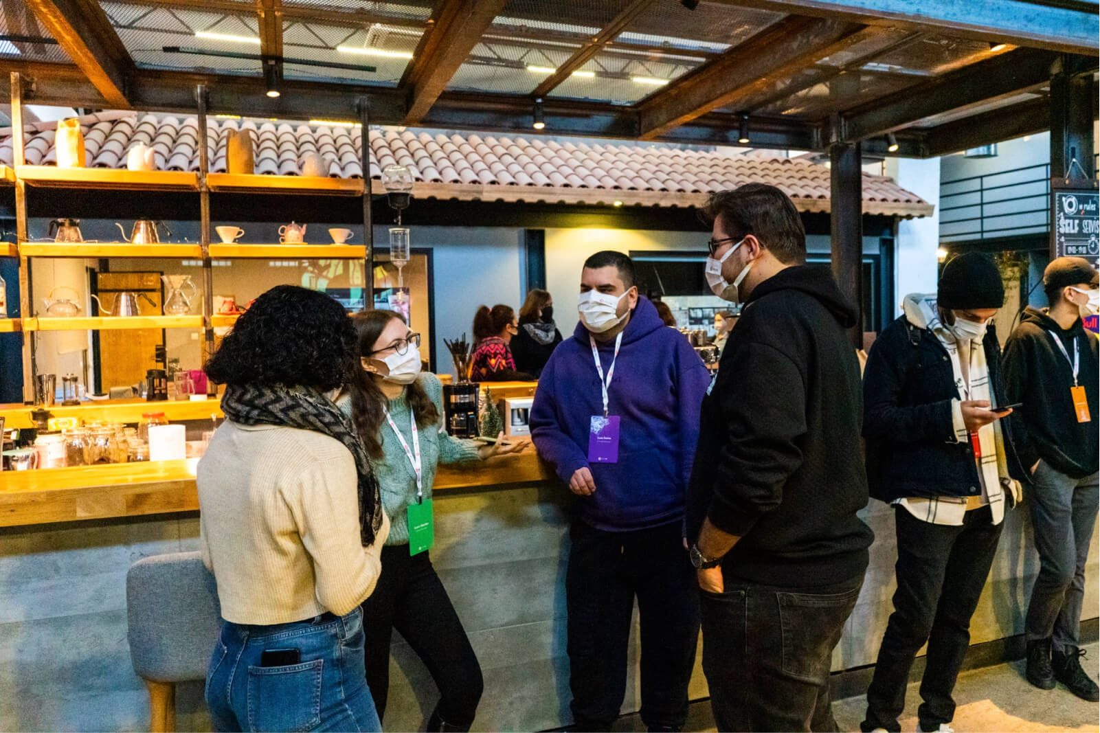 A group of people, standing in front of a coffee counter, wearing face masks, and talking to each other.