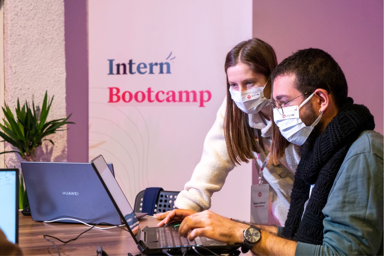 A man and a woman, wearing white face masks and looking at a laptop in front of a banner that says “Intern Bootcamp.”