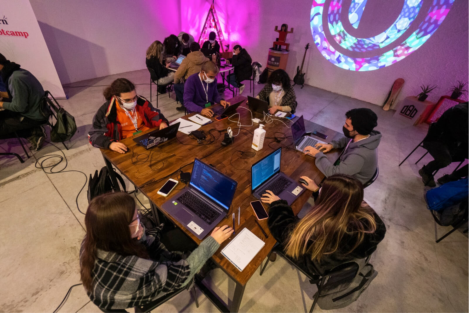 A group of people sitting around a wooden table working on their laptops and wearing face masks.