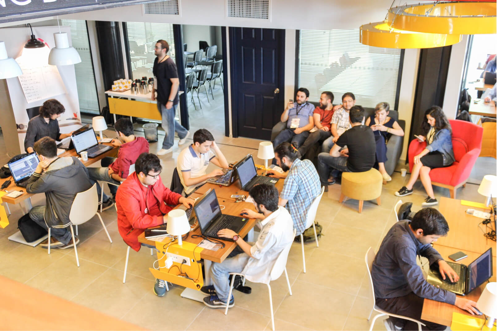 A group of people sitting at wooden desks, working on their laptops, and people sitting on a couch having a conversation.