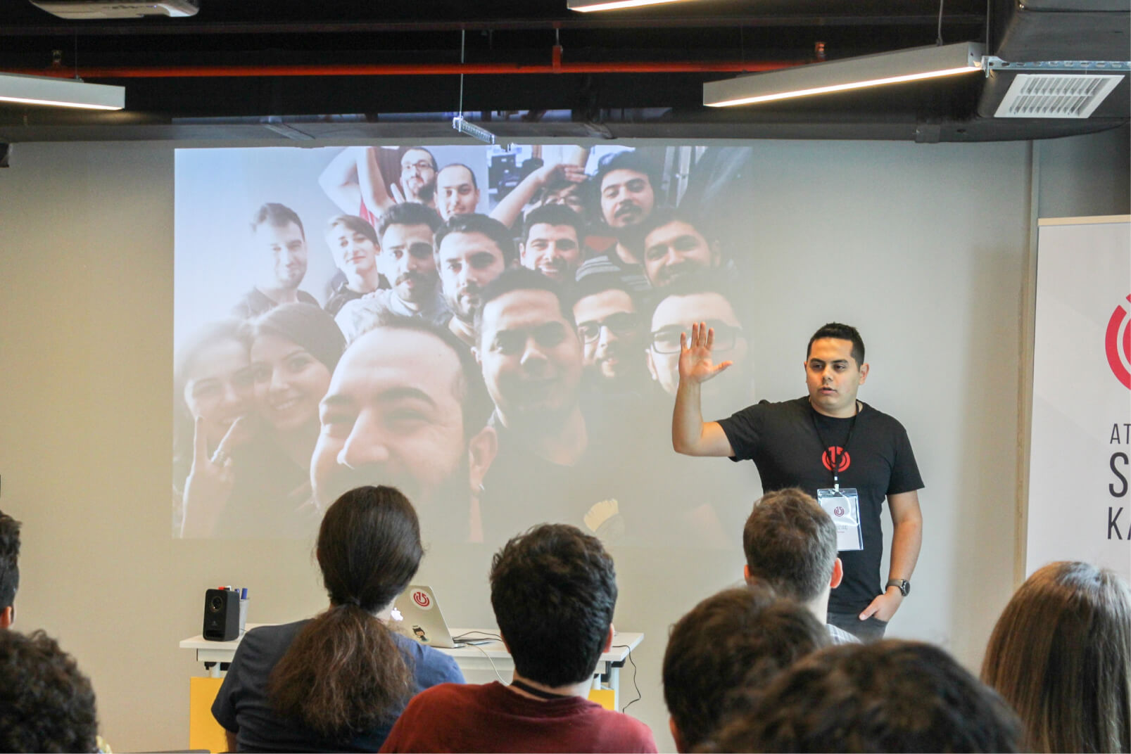 A man in a black shirt talking to people in front of a screen that shows Atolye15 team at the 1st Atolye15 Intern Bootcamp.