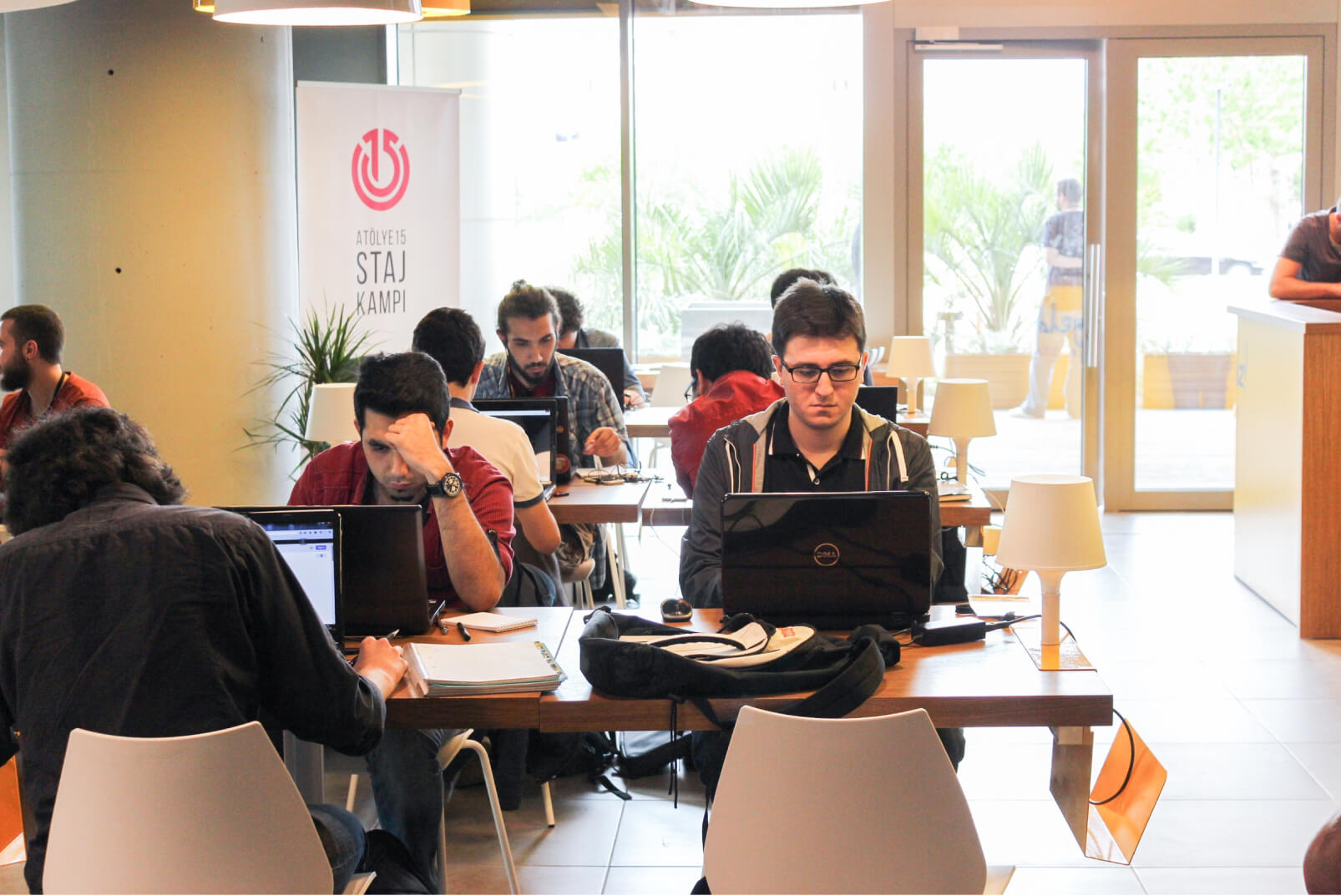 A group of young men sitting at desks, working on their laptops in front of a white banner that reads “Atölye15 Staj Kampı.”