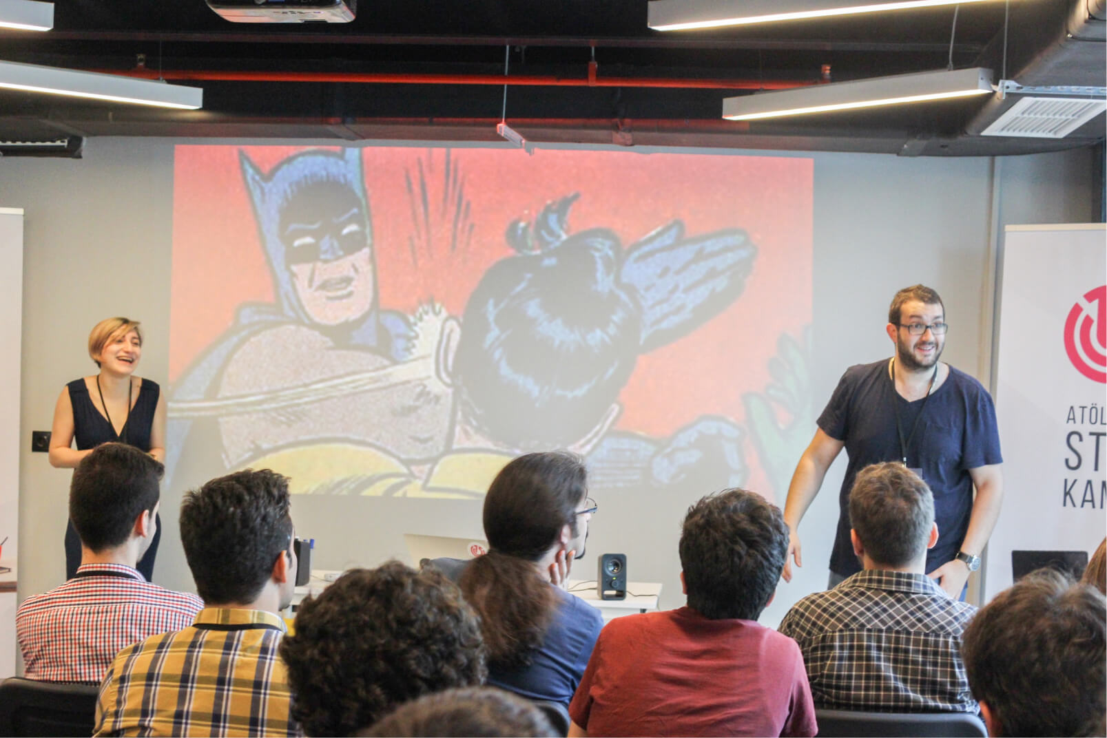 A man and a woman in front of a screen showing the “Batman slapping Robin” meme in front of a crowd at Atolye15 Intern Bootcamp.