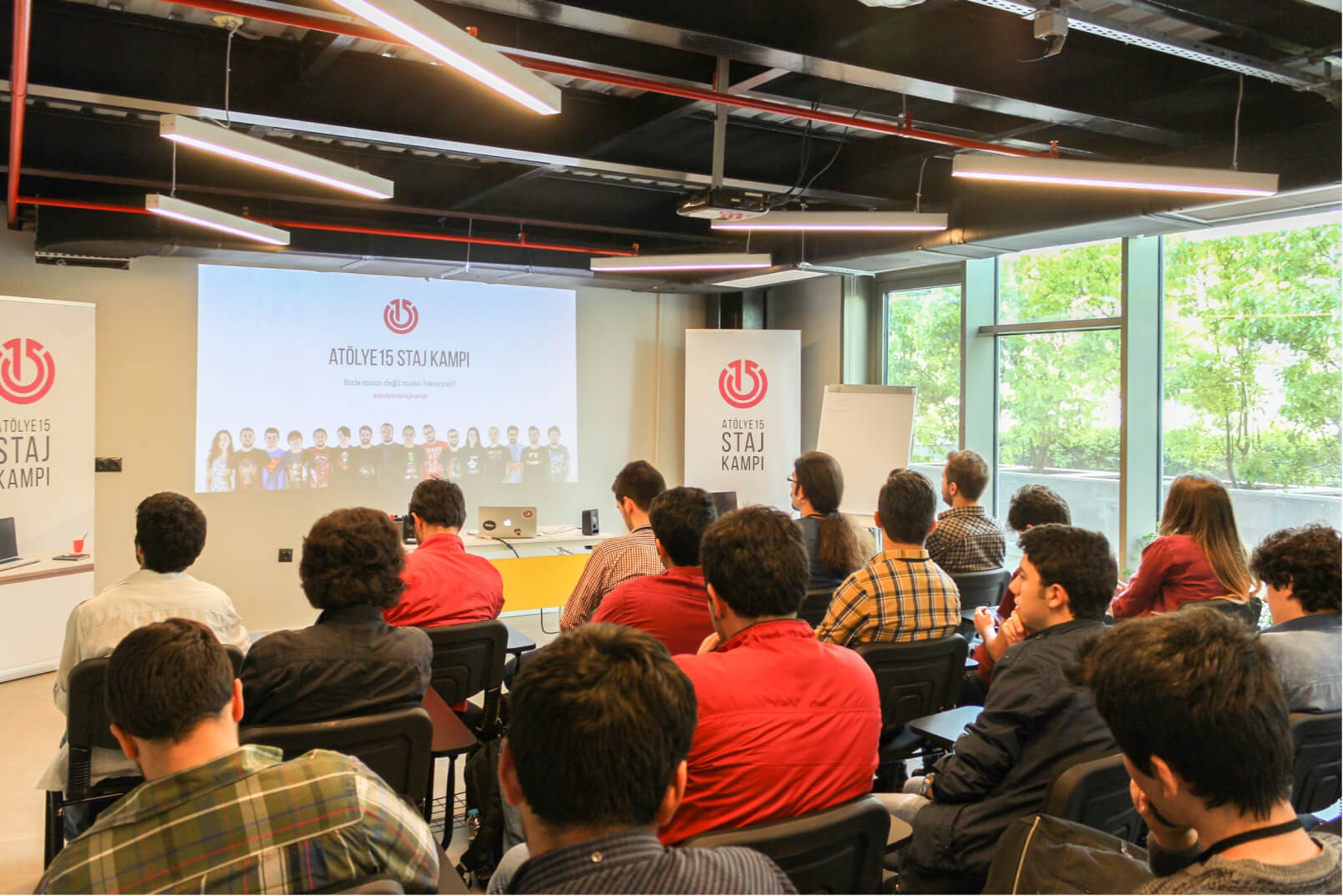 A group of people sitting in a conference room, looking at a screen that reads “Atölye15 Staj Kampı.”