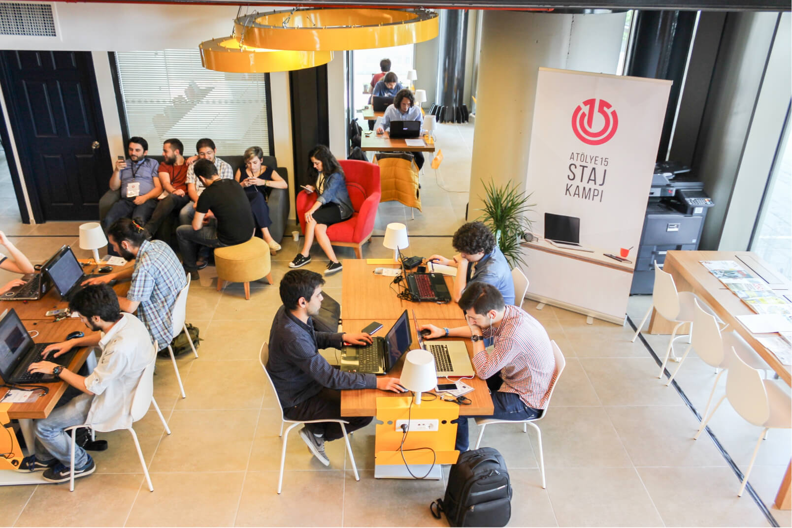 A group of people sitting at wooden desks, working on their laptops in front of a white banner that reads “Atölye15 Staj Kampı.”