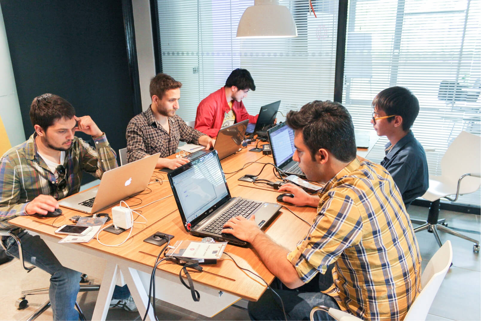 A group of young men sitting around a wooden desk, working on their laptops at the first Atolye15 Intern Bootcamp.