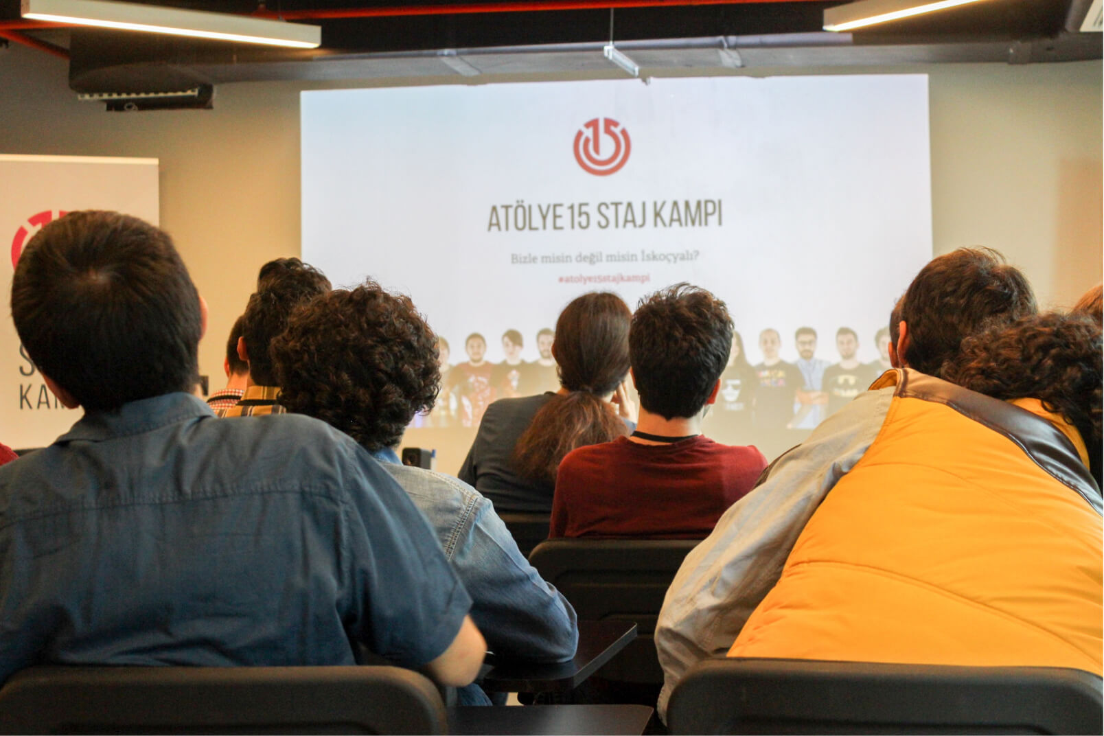 A group of people sitting in a conference room, looking at a screen that reads “Atölye15 Staj Kampı.”