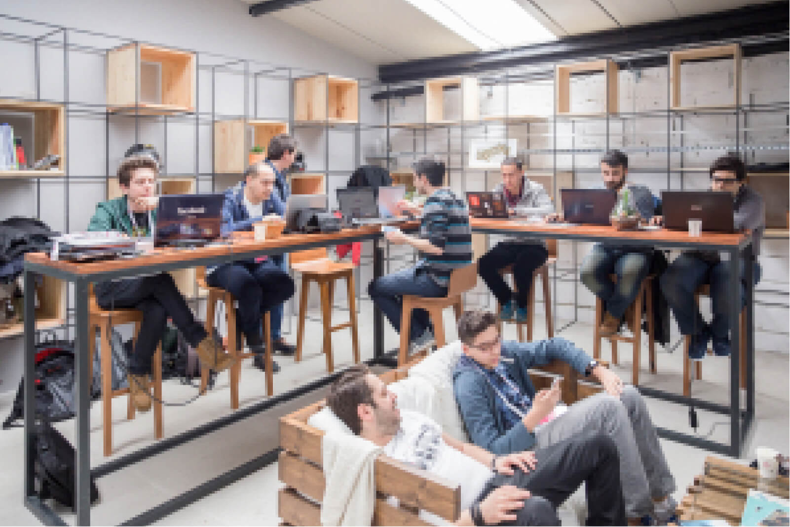 A group of young people sitting on counter stools at a wooden desk, working on their laptops in front of a bookshelf.