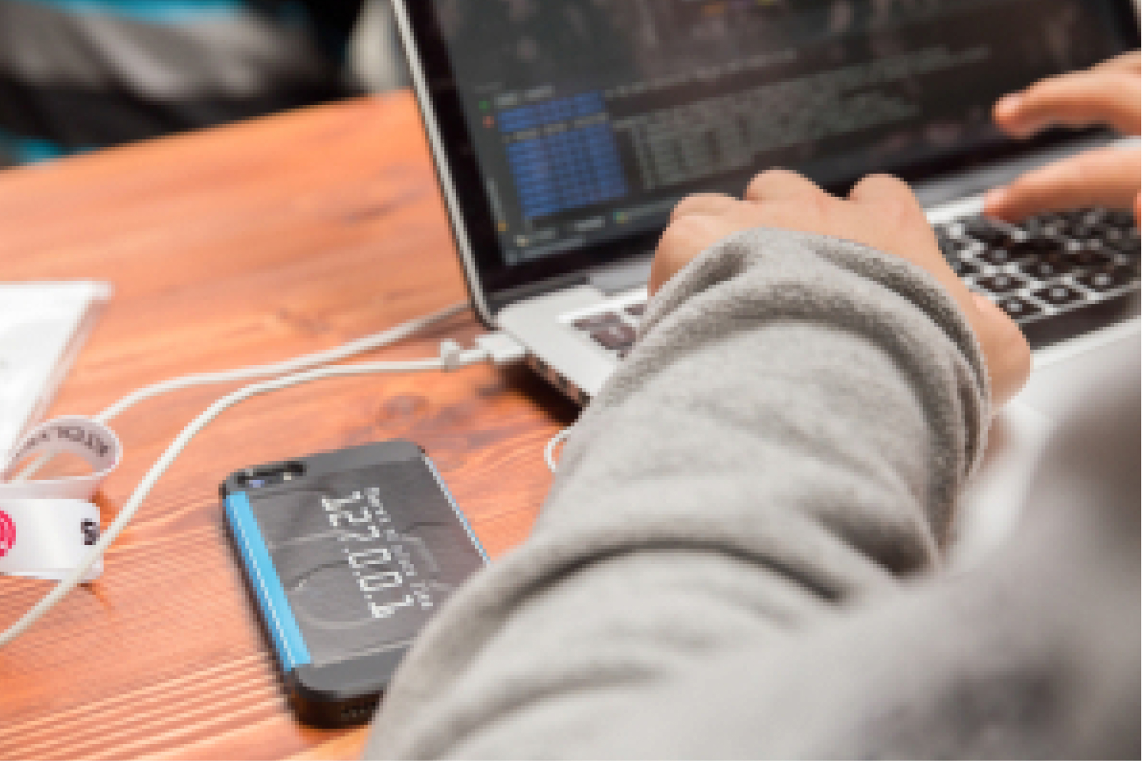 Hands in a long-sleeved grey sweatshirt, typing on a keyboard, a MacBook screen showing lines of code, and a phone on a table.