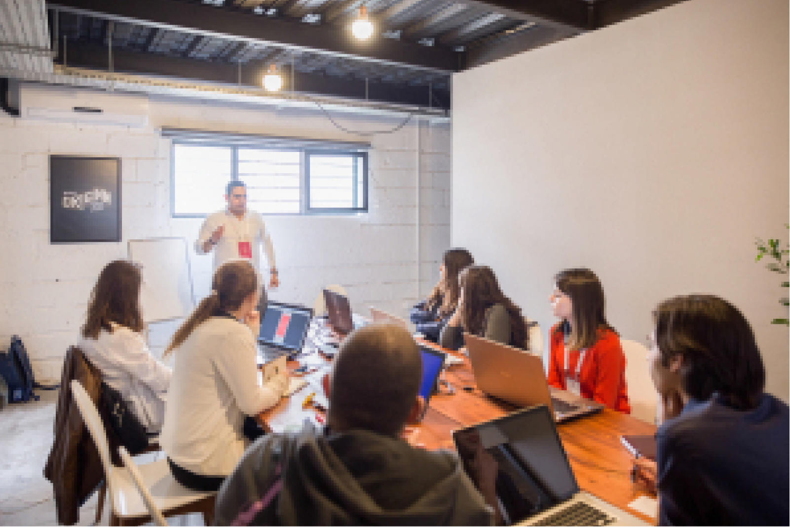 A man in a white shirt talking to a group of people sitting around a table with their laptops in a white room.