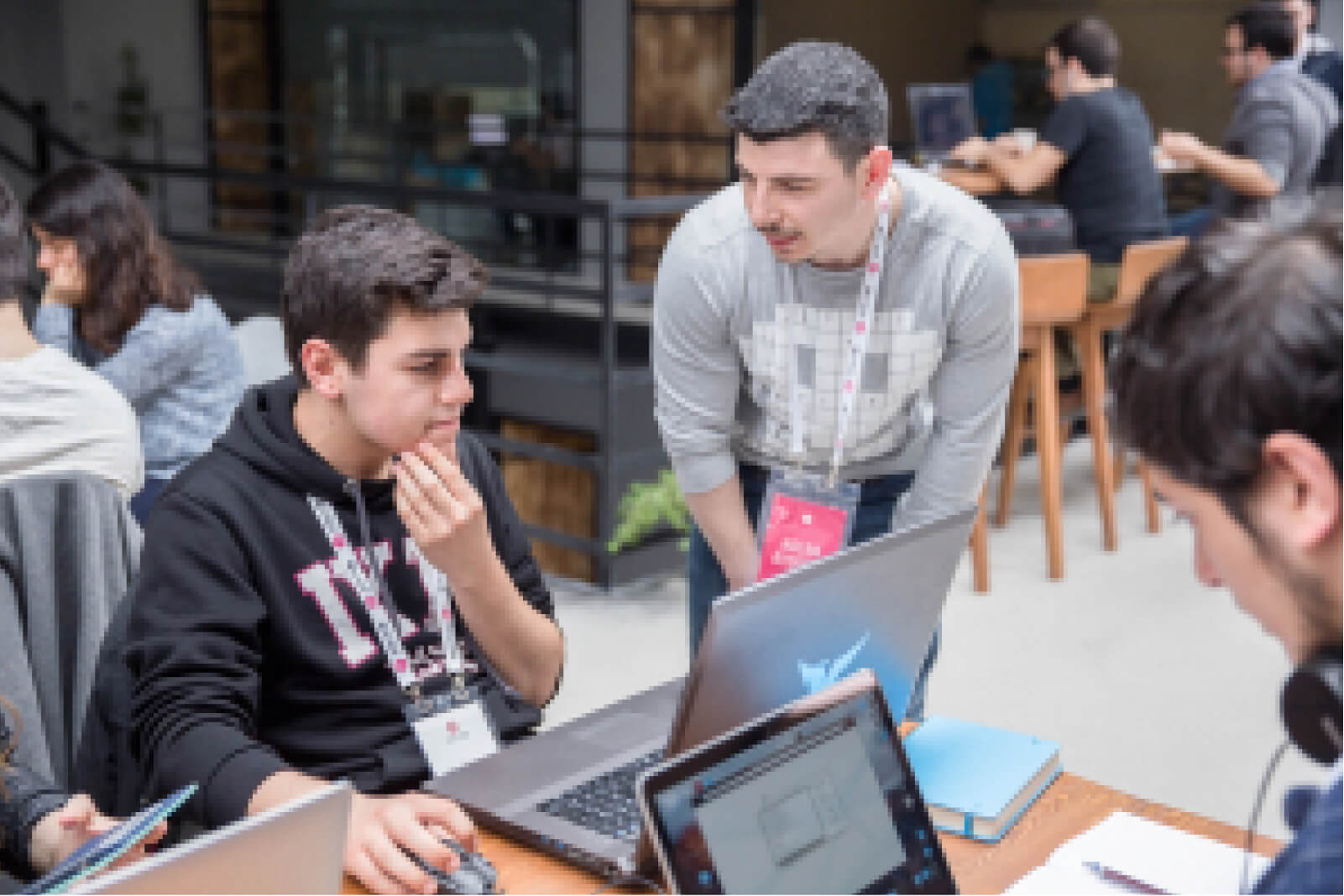 A man in a grey sweatshirt slightly bent over, talking to a young bot in a black hoodie, holding his chin and using a laptop.