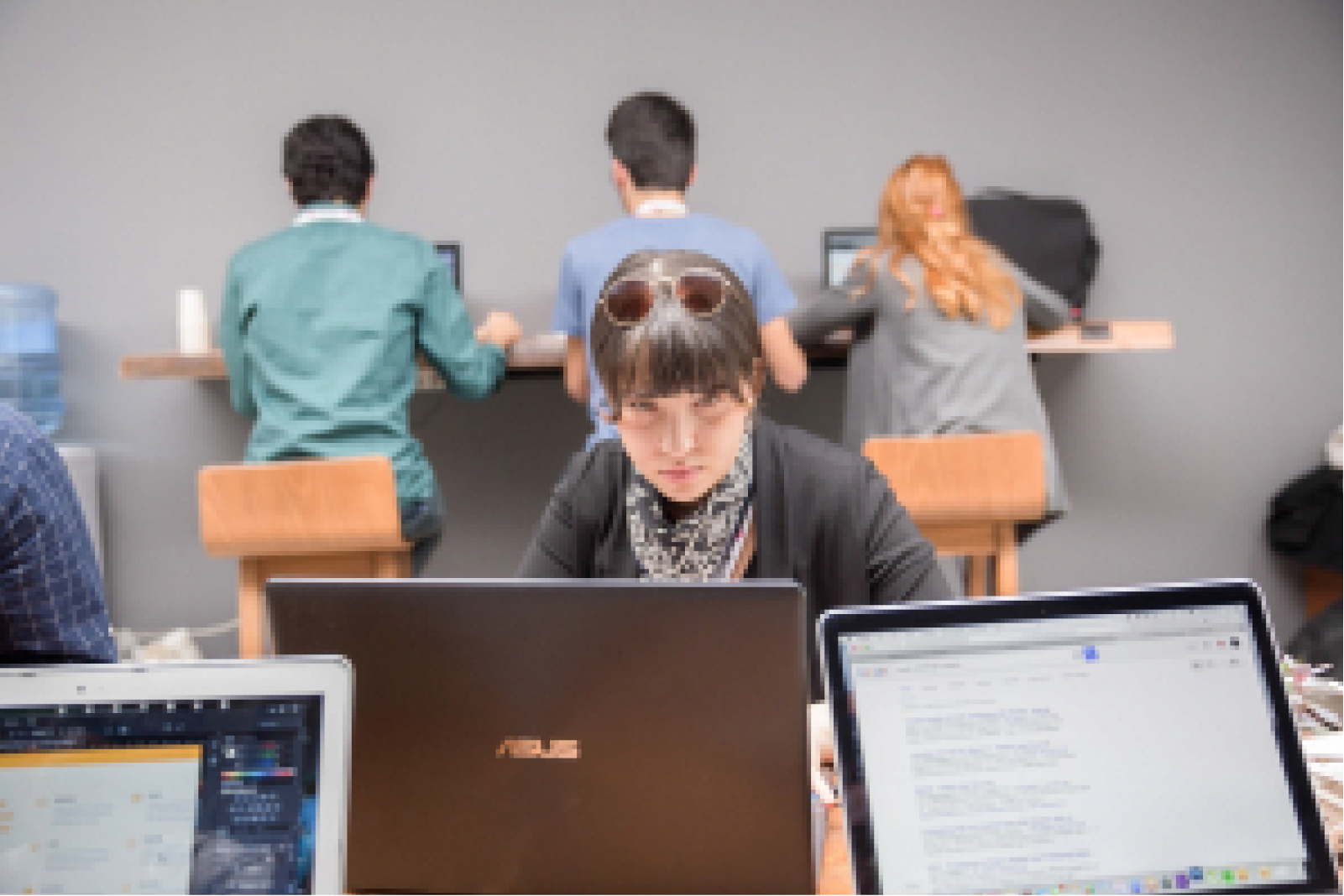 A young woman with bangs, wearing a grey scarf and sunglasses on her head, looking at a laptop and 3 people in the back.