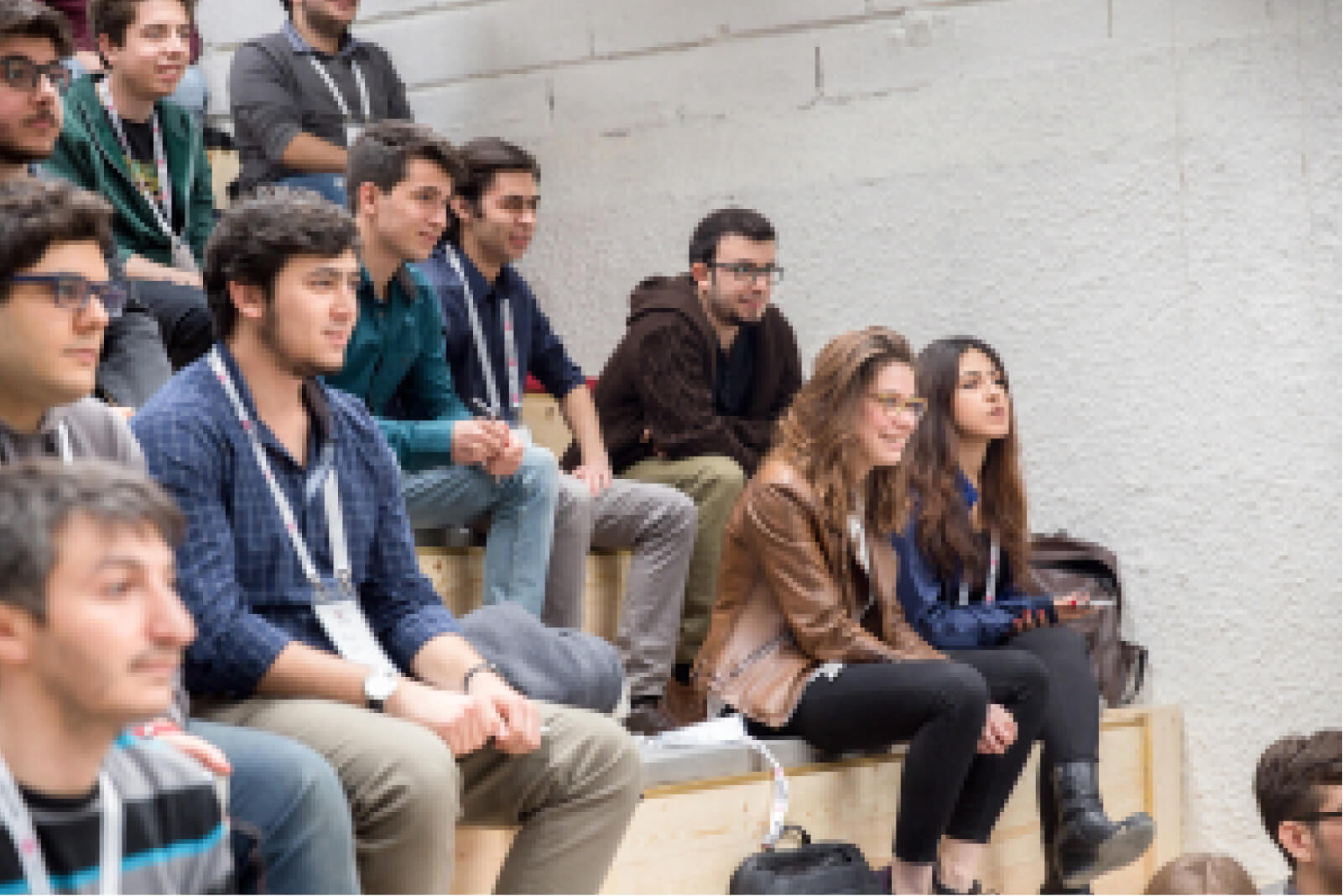 A group of young people sitting in a lecture hall, carefully listening to a speech next to a white brick wall.