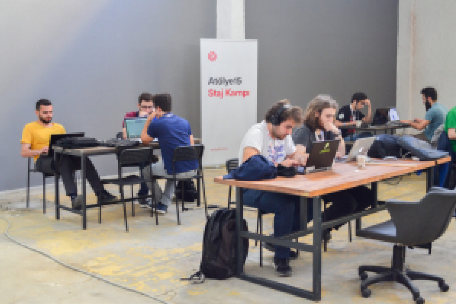 A group of young people, sitting at tables and working on laptops in front of a white banner that says “Atölye15 Staj Kampı.”