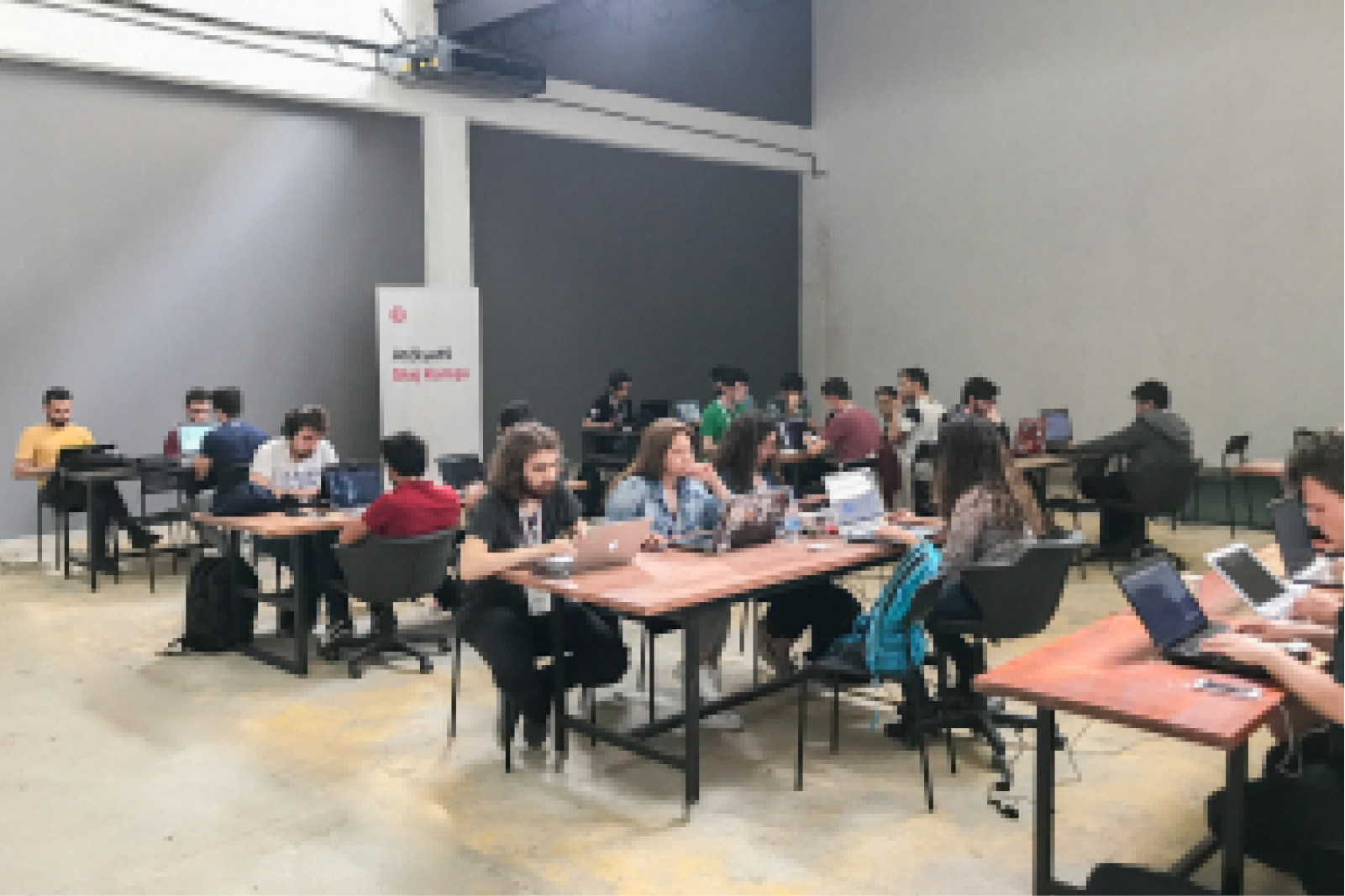 A group of young people, sitting at tables and working on laptops in front of a white banner that says “Atölye15 Staj Kampı."