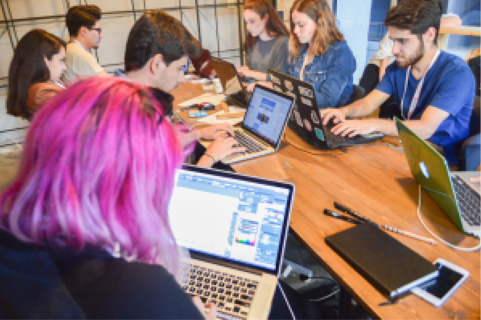 A girl with pink hair using a MacBook, sitting among other young people working on their laptops at a desk.