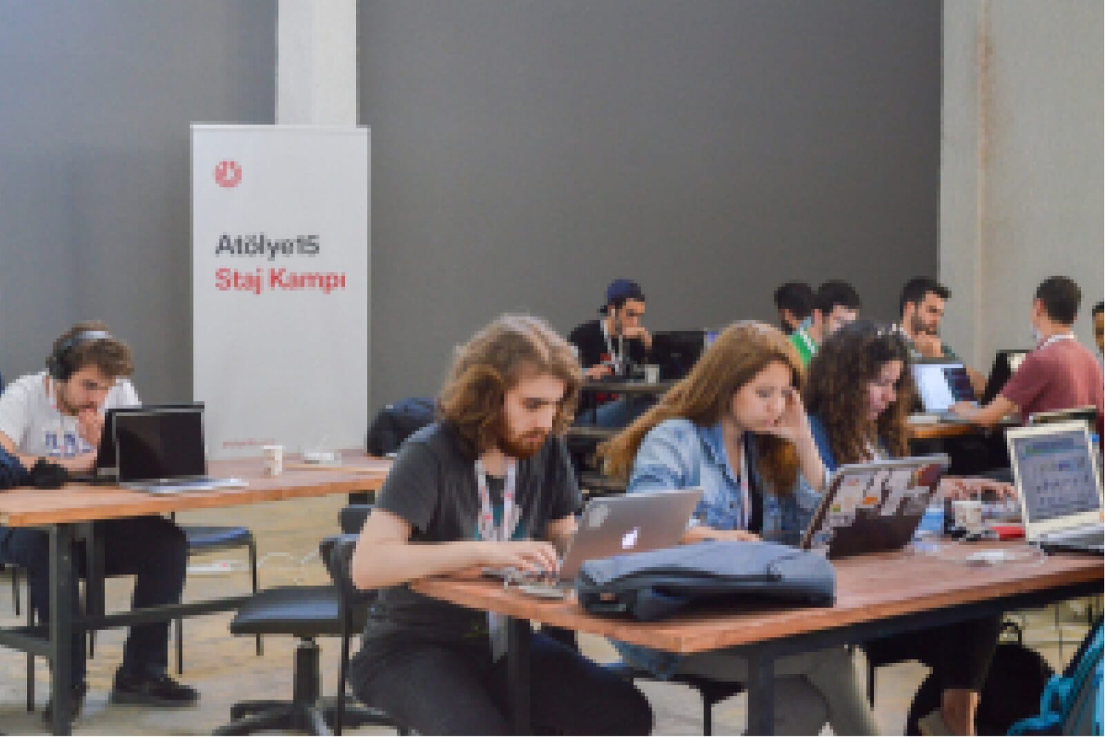 A group of young people, sitting at tables and working on laptops in front of a white banner that says “Atölye15 Staj Kampı.”