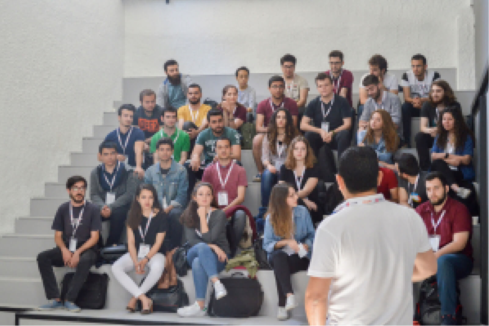 A man in a white t-shirt with his back turned to the camera, standing in front of people sitting in a lecture hall.