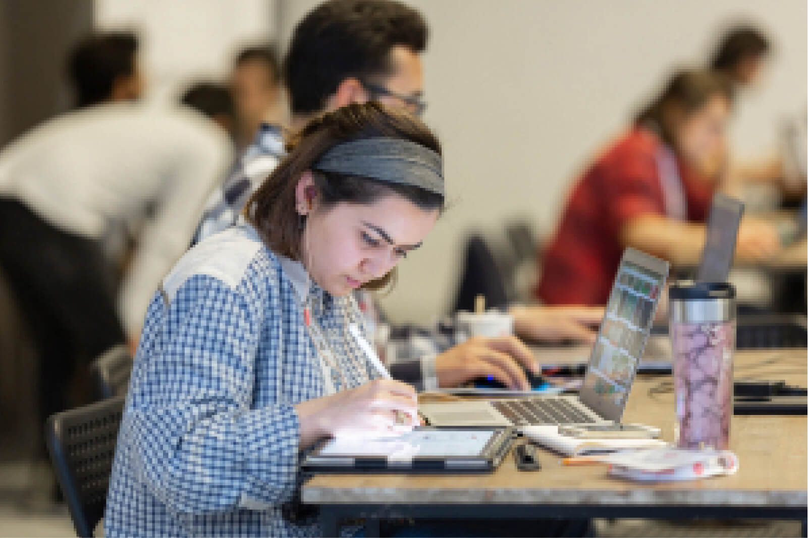 A young girl in a plaid shirt, wearing headphones and a headband, sitting in front of a laptop, drawing on a tablet.
