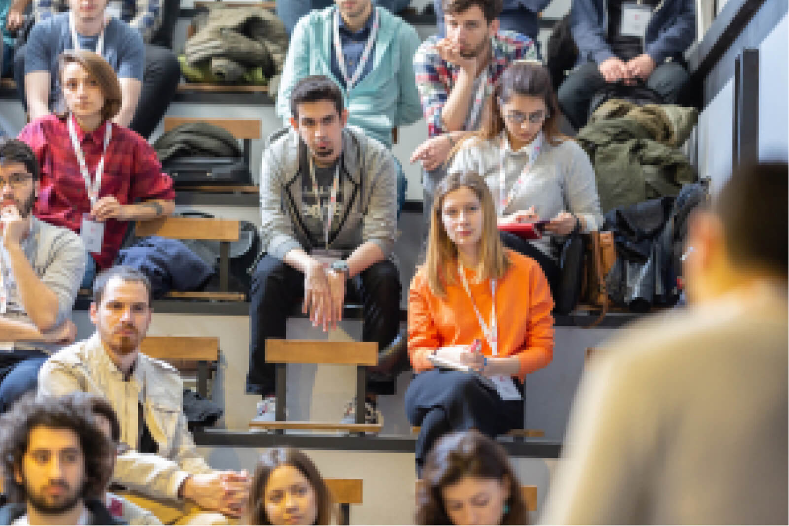 A blond girl in an orange sweatshirt among a group of people sitting in a lecture hall and looking at a man on stage.