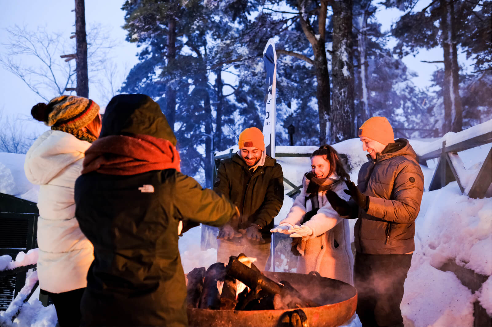 A group of people in winter coats and hats warming themselves by a fire pit filled with wood surrounded by trees and snow.