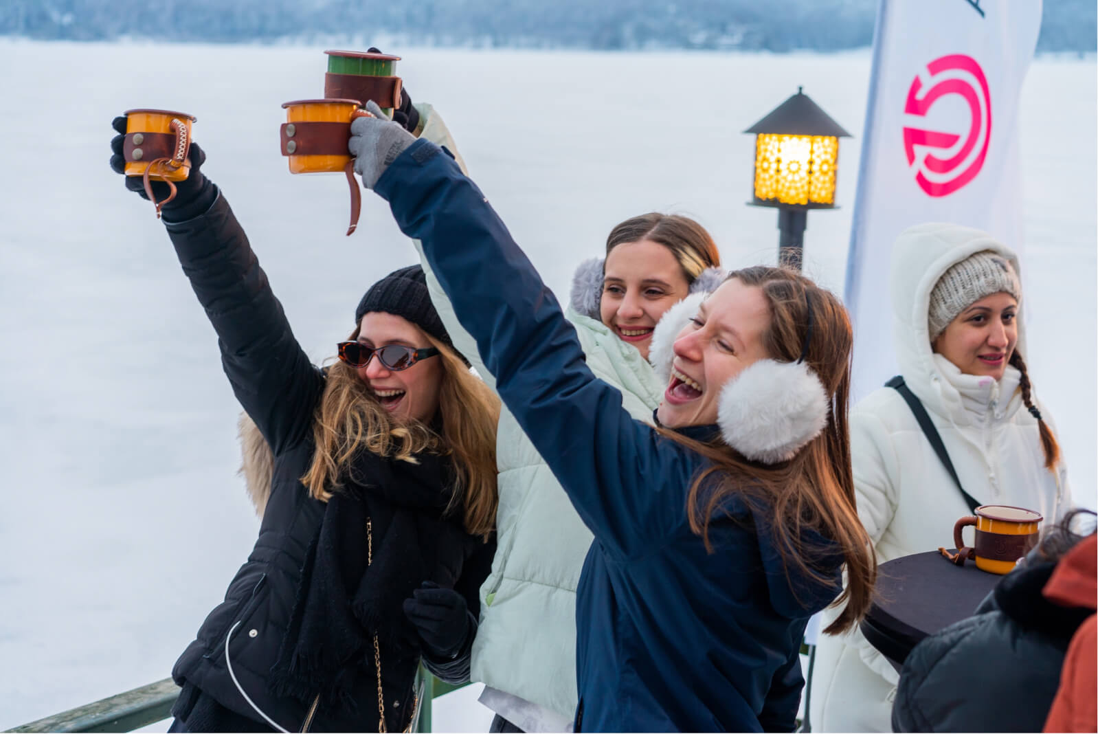 4 women wearing winter coats and earmuffs, raising their cups in the air, a lamp, and the Atolye15 logo surrounded by snow.