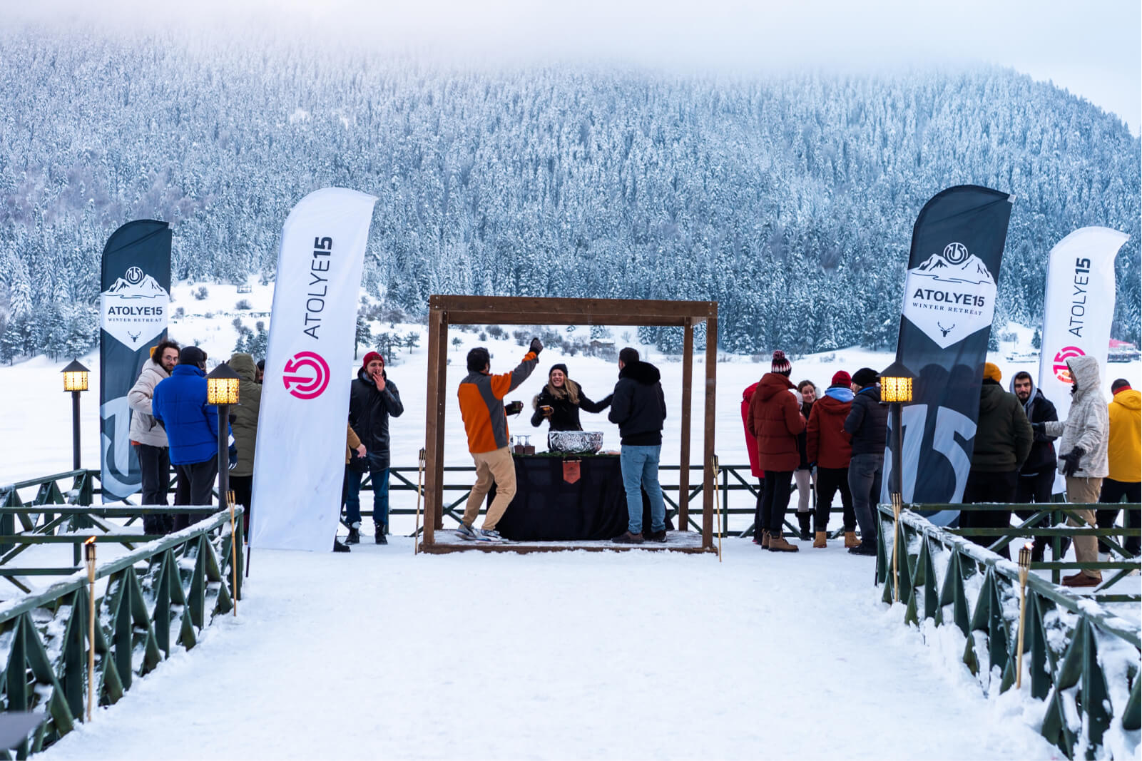 A group of people in winter coats on a snow-covered deck by a snowy mountain and the Atolye15 logo and Winter Retreat banners.