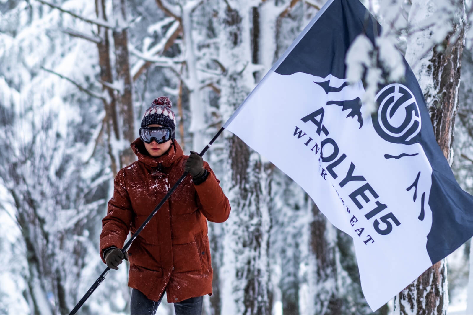A man in a brown winter coat wearing ski goggles and black gloves holding a flag that says Atolye15 Winter Retreat.