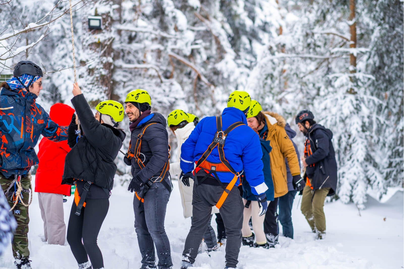 A group of people in the snow, wearing winter coats, helmets, and straps as zipline gear.