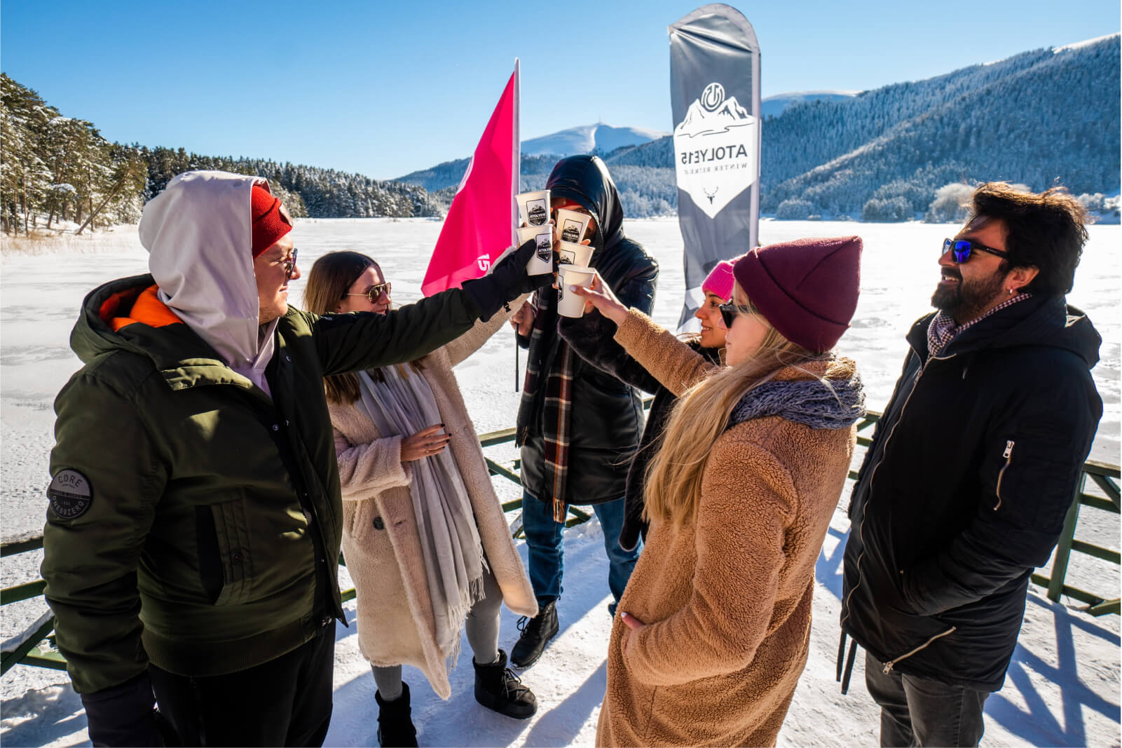 People in winter coats and hats raising their glasses surrounded by snow in front of an Atolye15 Winter Retreat banner.