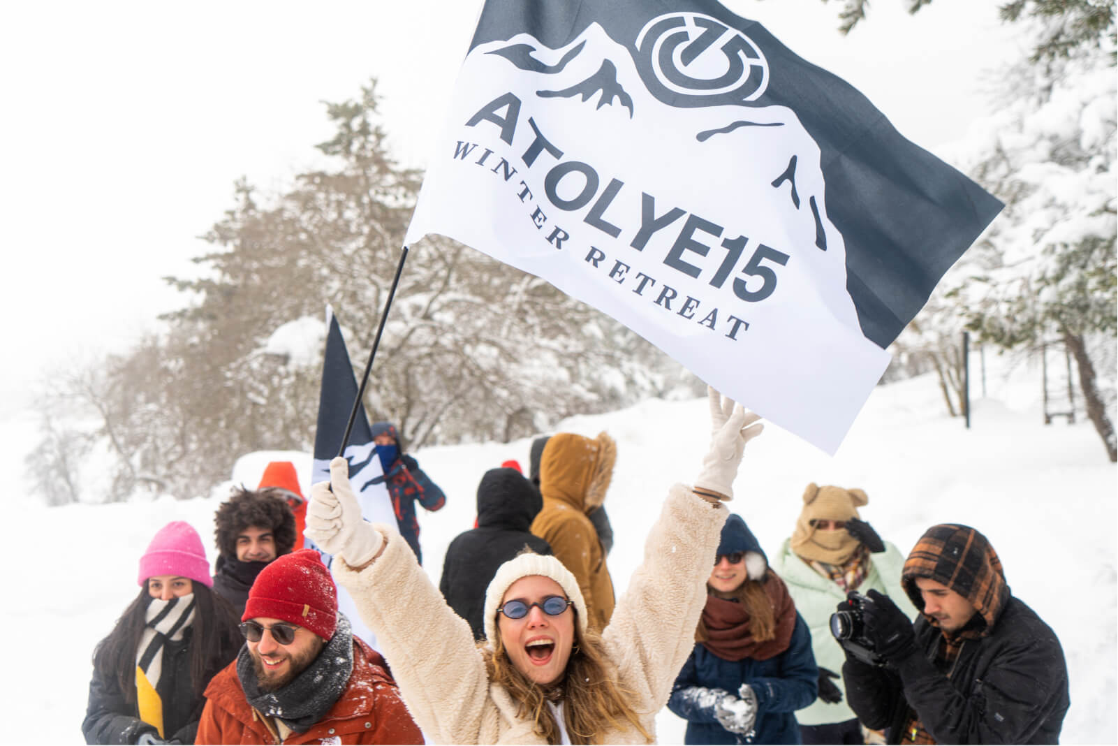A woman wearing sunglasses, gloves, and a white hat in front of people surrounded by snow and the Atolye15 Winter Retreat flag.