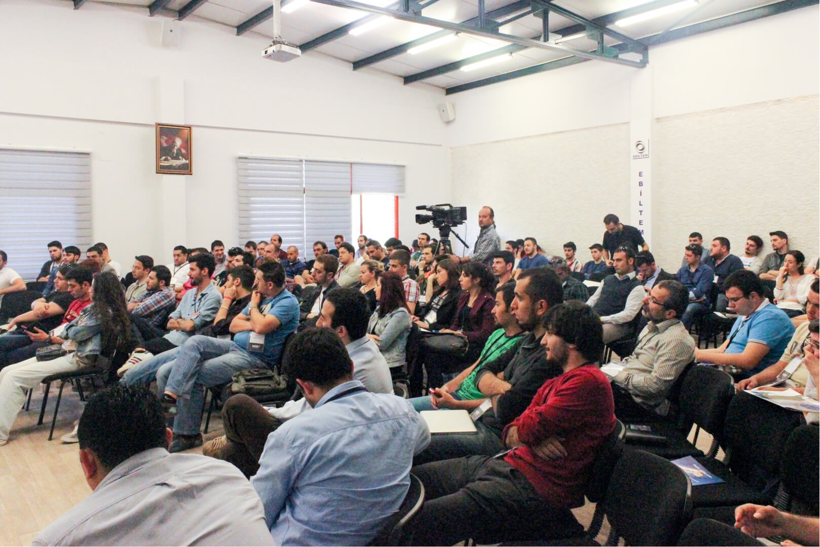 A crowd of people sitting in a white conference room and a man with a camera recording.