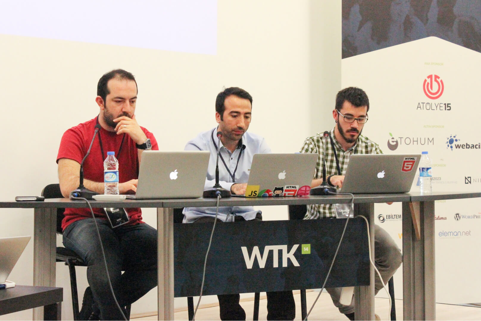 Three men, sitting at a WTK table using their MacBooks in front of a banner filled with logos including the Atolye15 logo.
