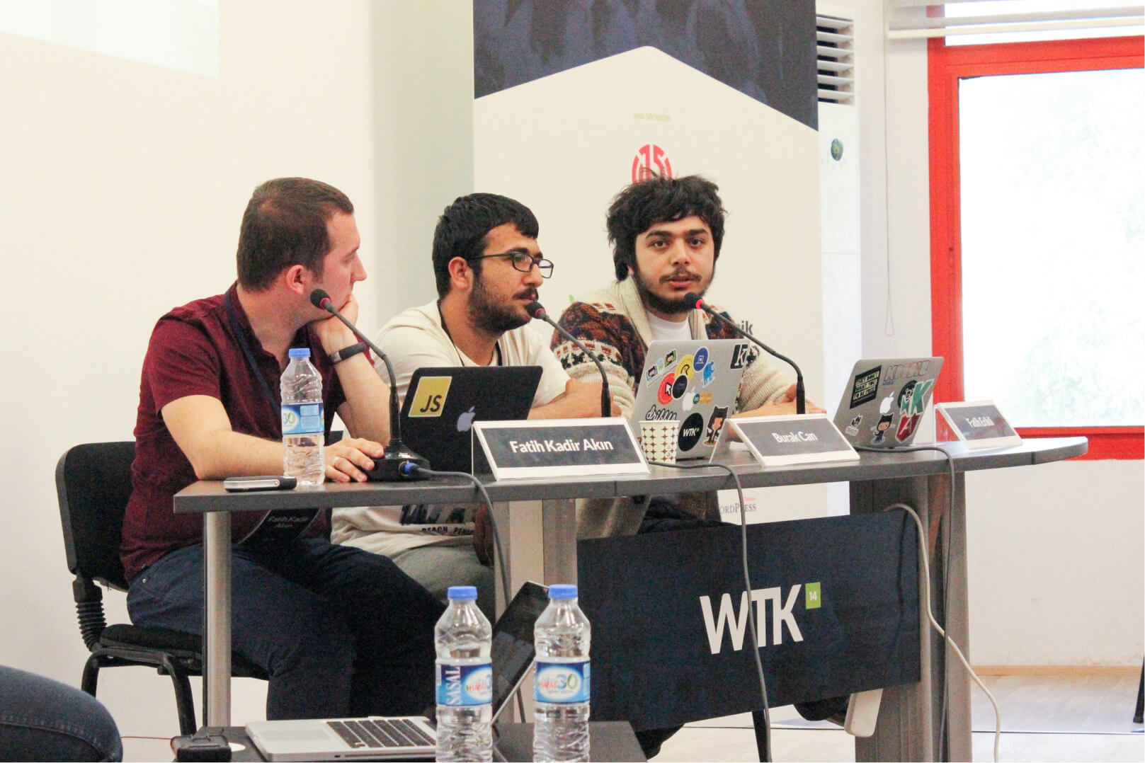 Three men, sitting at a WTK desk in front of their MacBooks, microphones, and a banner with the Atolye15 logo on it.