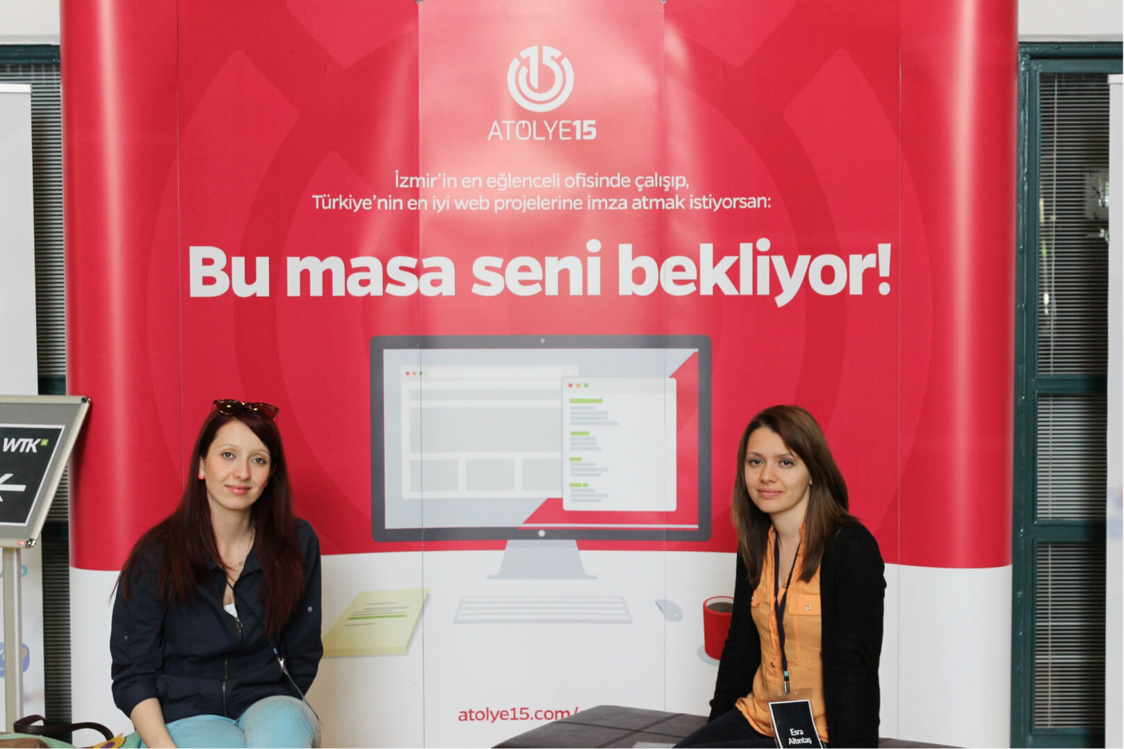 Two women, sitting and posing in front of a pink banner that reads “Bu masa seni bekliyor!” under the Atolye15 logo.