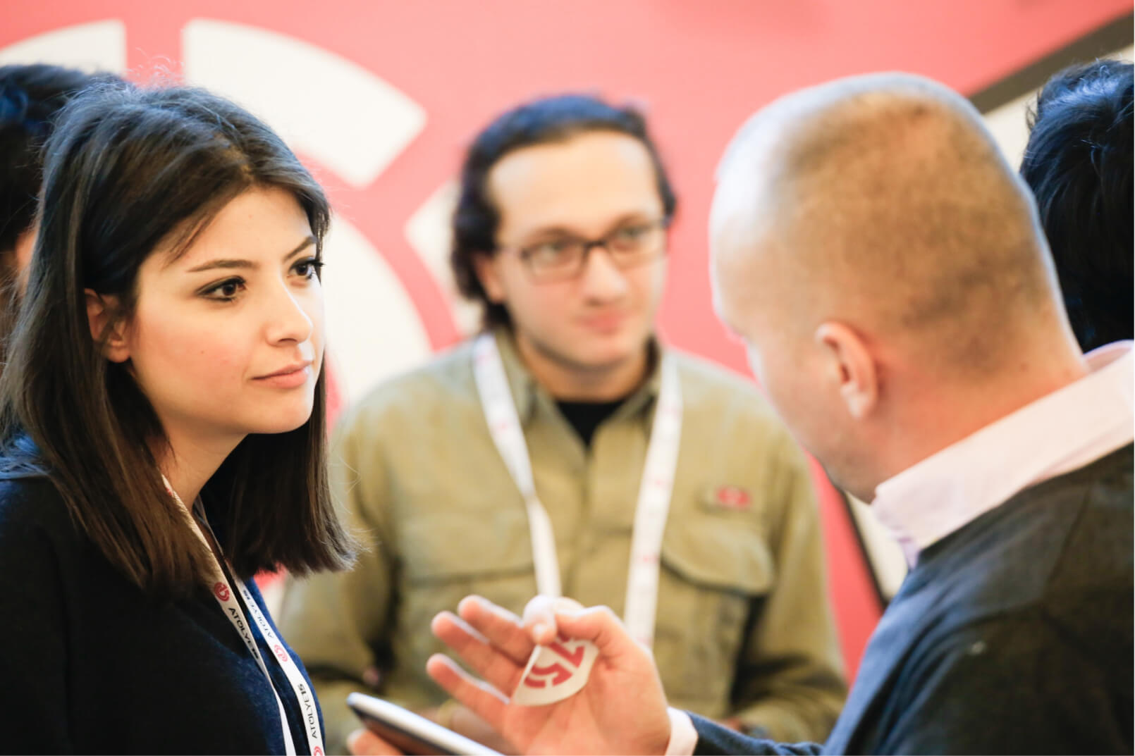 A man holding the Atolye15 logo, talking to a young brunette woman, and a man in a green shirt in the background.