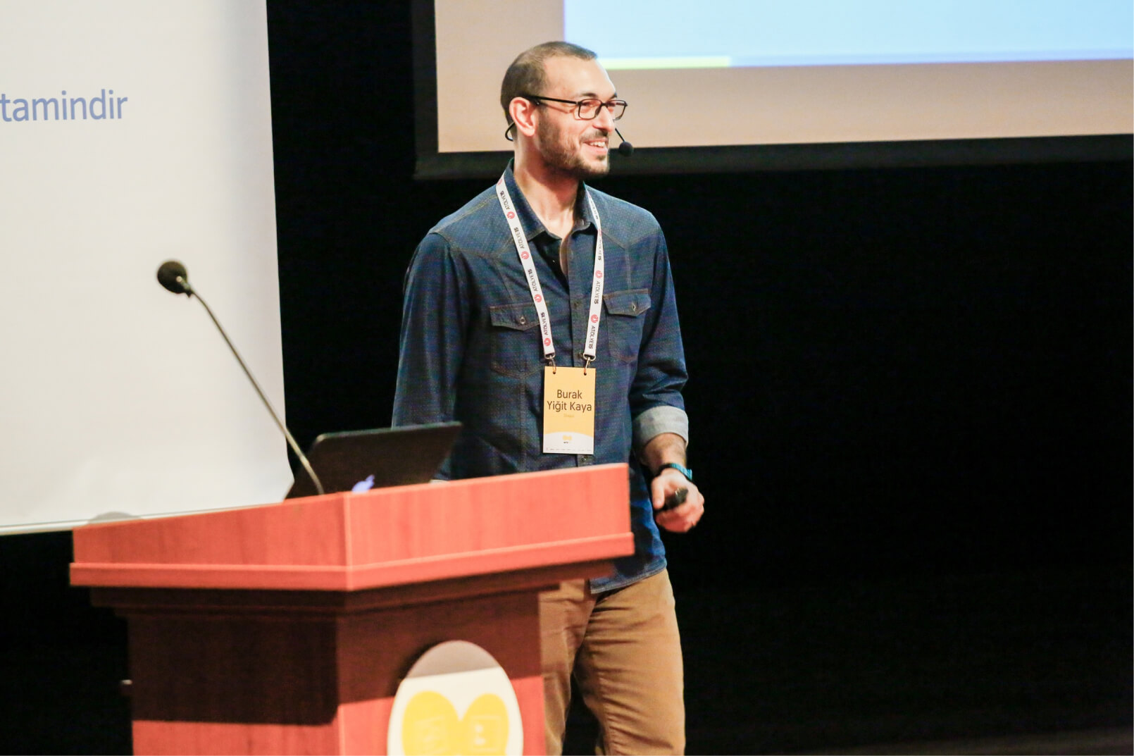 A man in a denim shirt, wearing glasses and a name tag that reads “Burak Yiğit Kaya” giving a speech next to a lectern.
