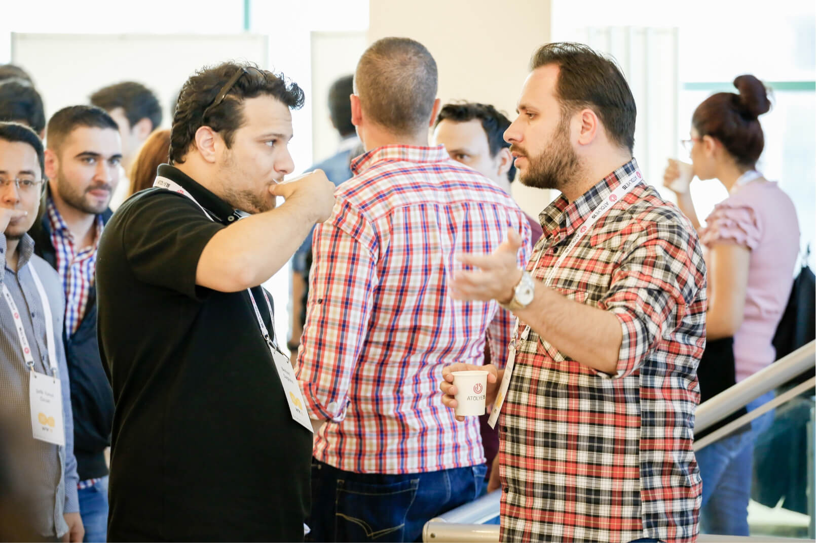 A man in a black shirt drinking coffee, talking to a man in a plaid shirt holding a carton cup in front of a crowded background.