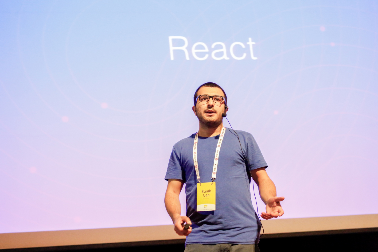 A man in a blue t-shirt, wearing glasses, a microphone, and a name tag that reads “Burak Can” in front of a screen saying React.