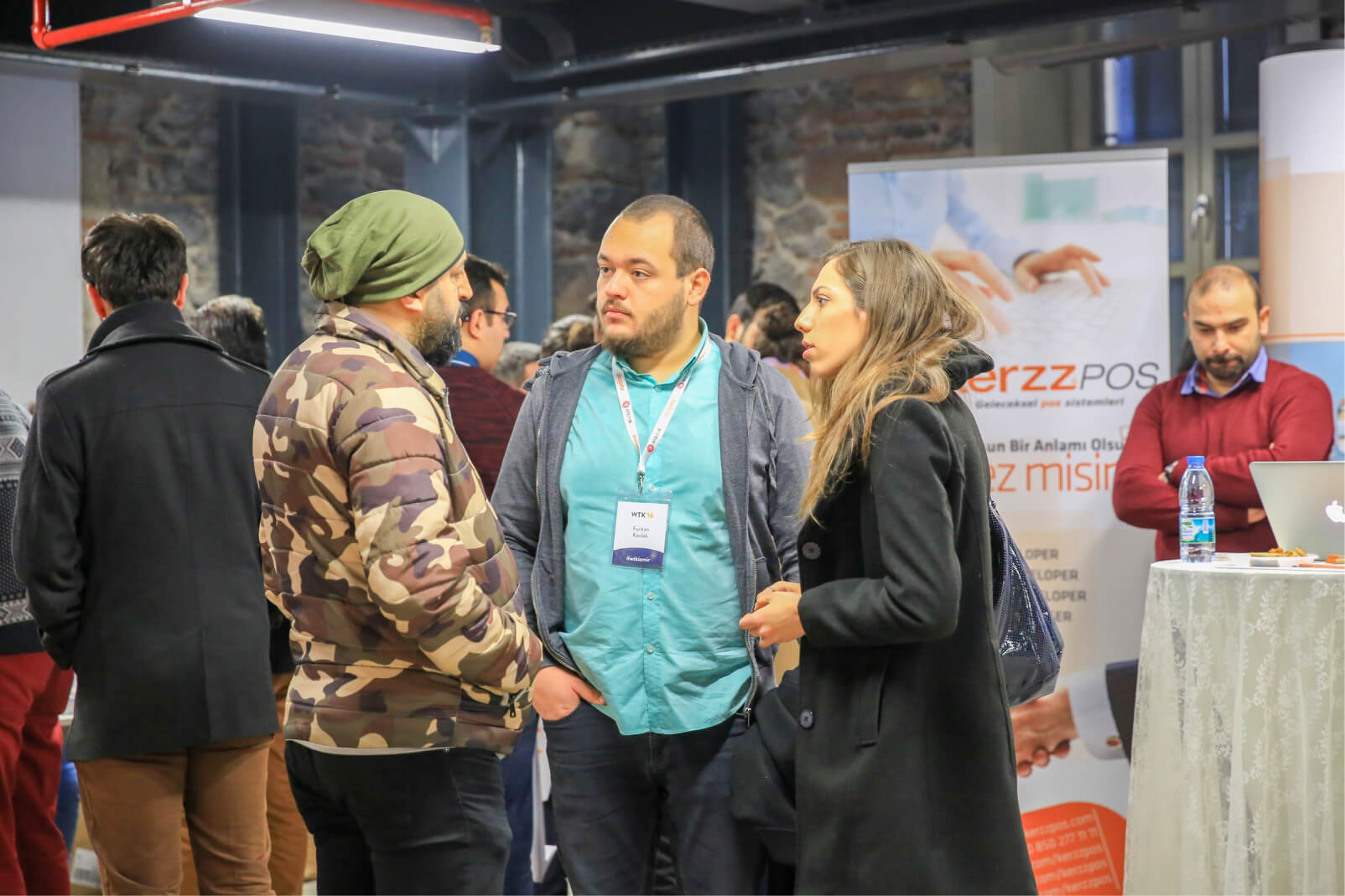 A man in a green hat and a camouflage jacket, a man in a blue shirt, and a long-haired woman in a black overcoat talking.