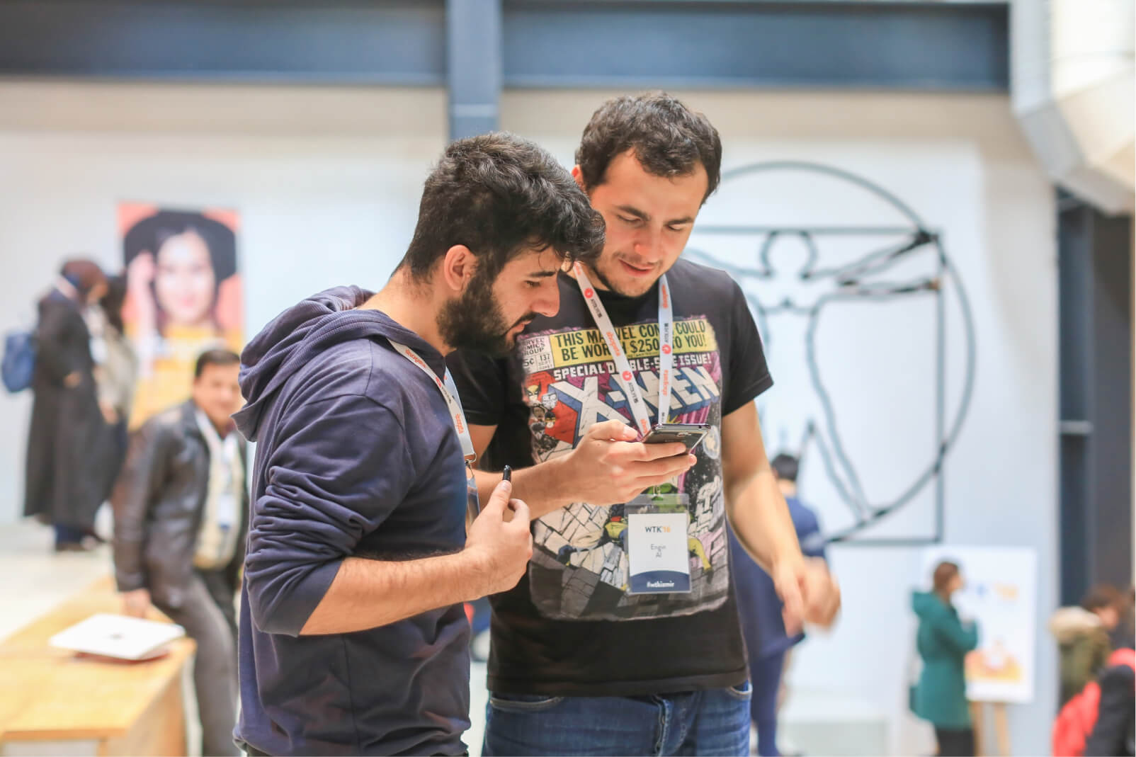 A young bearded man in a hoodie and another man in an X-Men t-shirt looking at a smartphone together.