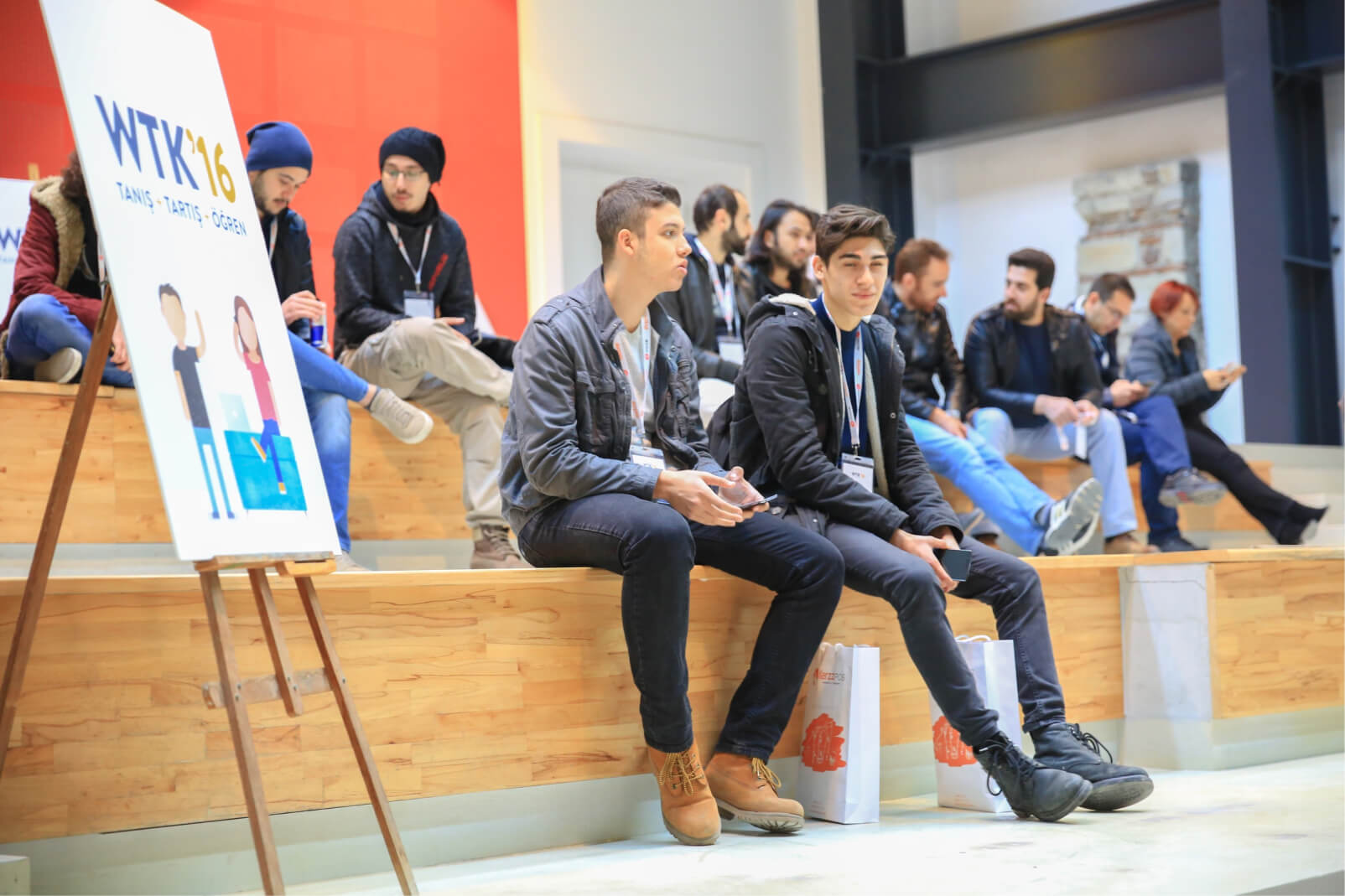 A group of young people talking while sitting on wooden stairs next to a whiteboard that reads “WTK’16 Tanış, Tartış, Öğren.”