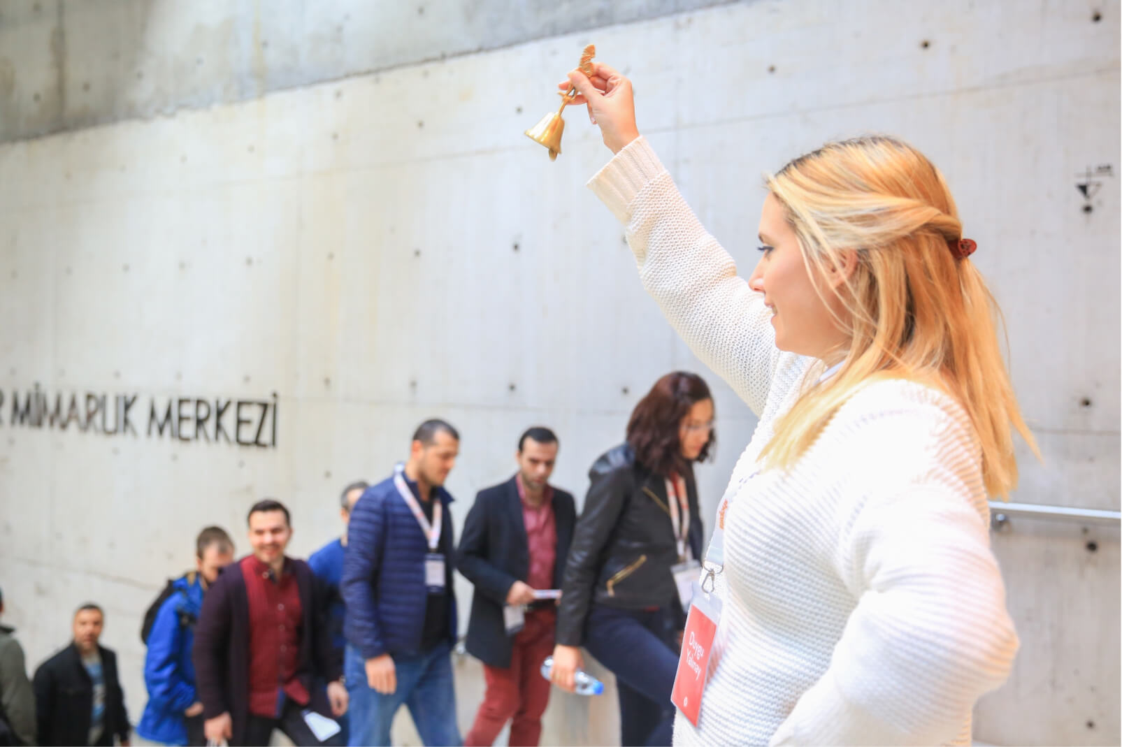 A blond woman in a white sweater, ringing a bell, and a line of people walking up the stairs by a wall saying “Mimarlık Merkezi.”