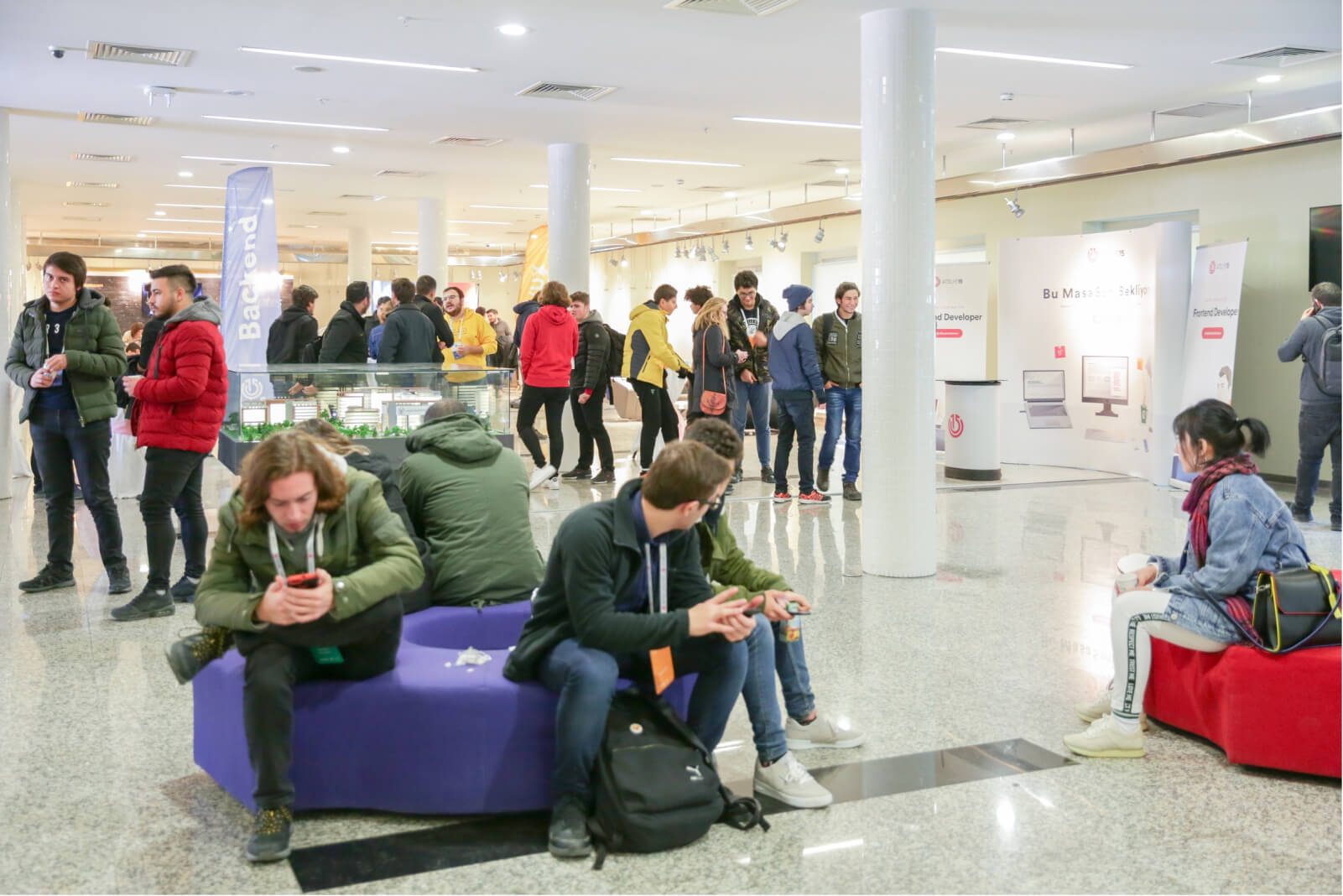 A group of people standing and sitting around talking and looking at their phones with Atolye15 WTK banners in the background.