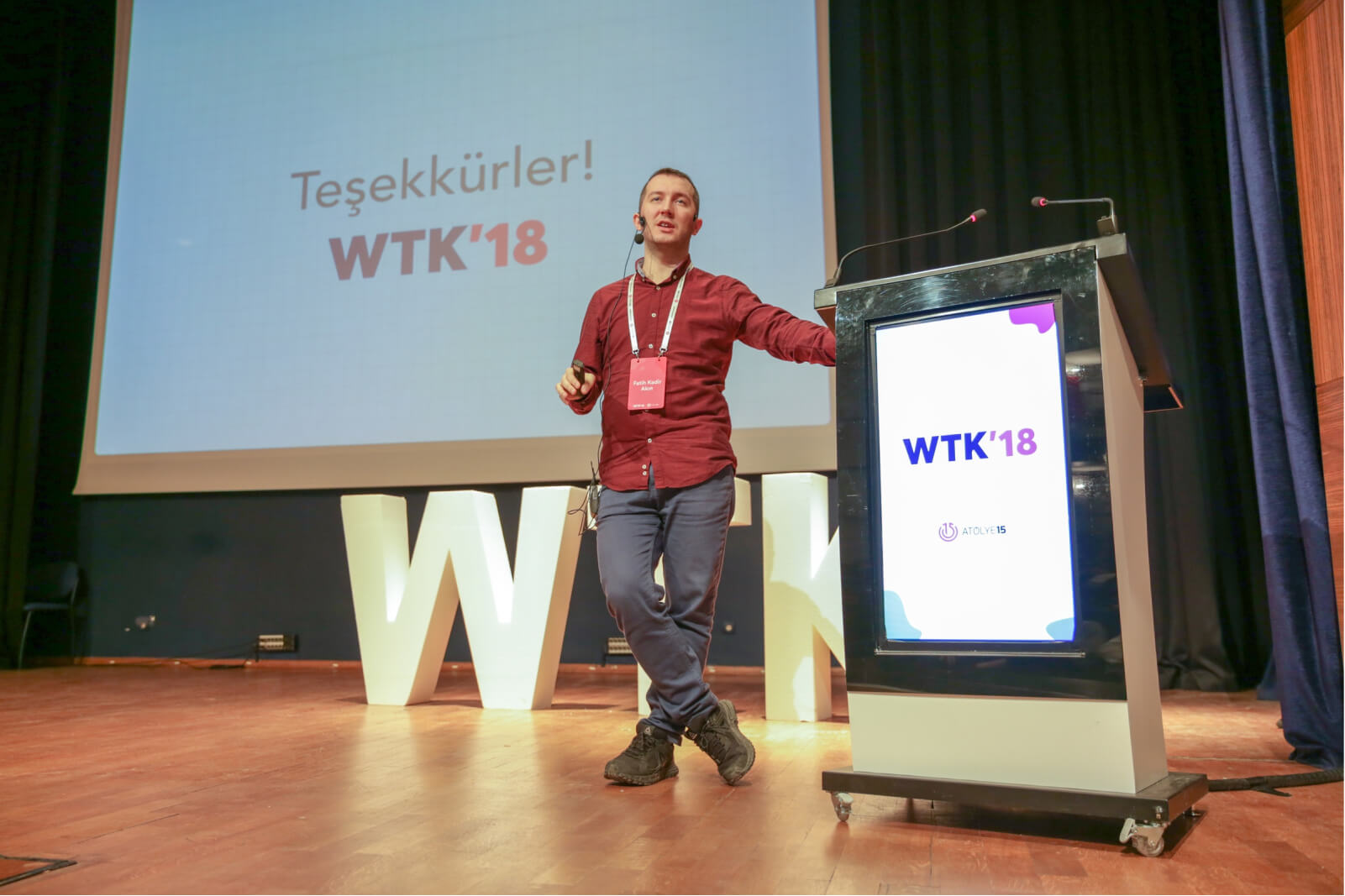 A man in a red shirt wearing a headset microphone, leaning against a lectern in front of a screen saying “Teşekkürler WTK’18."