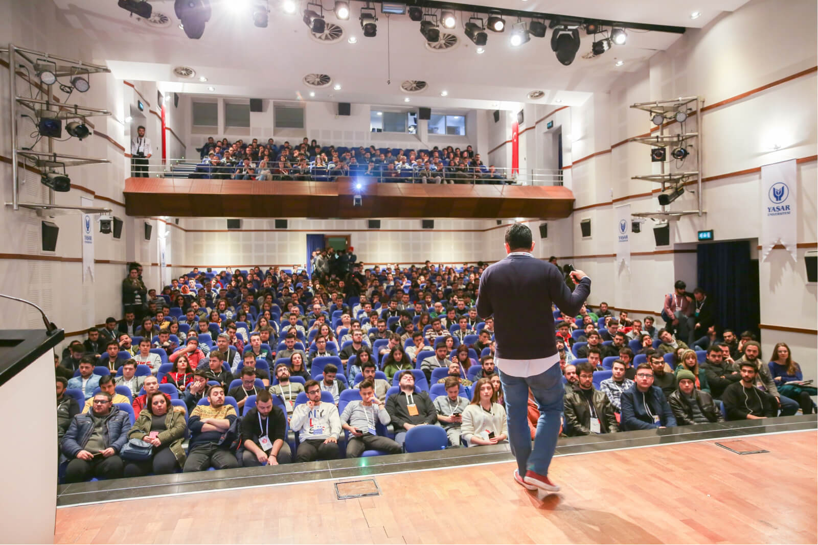 The back of a man on stage, speaking to a crowd of people in a conference hall, and the banner of Yaşar University on the wall.