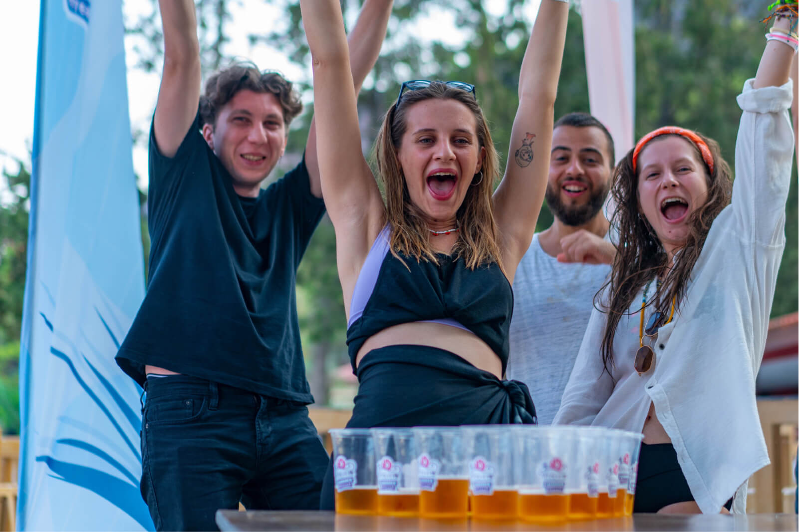 Two men and two women, cheering with their hands up in the air, playing beer pong in front of plastic beer glasses.