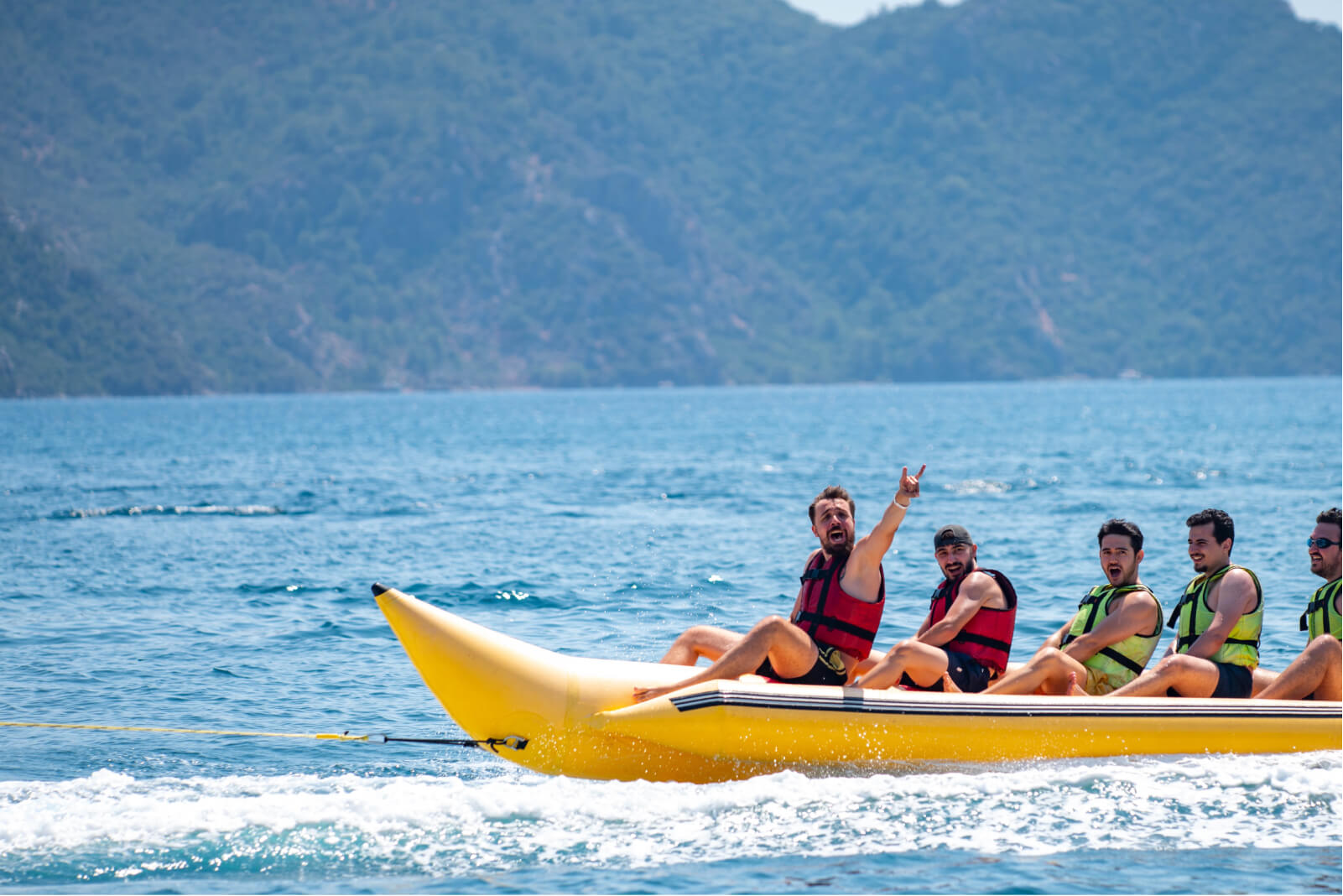 Five men on an inflatable yellow banana riding the waves and wearing life jackets in the sea by a mountain of trees.
